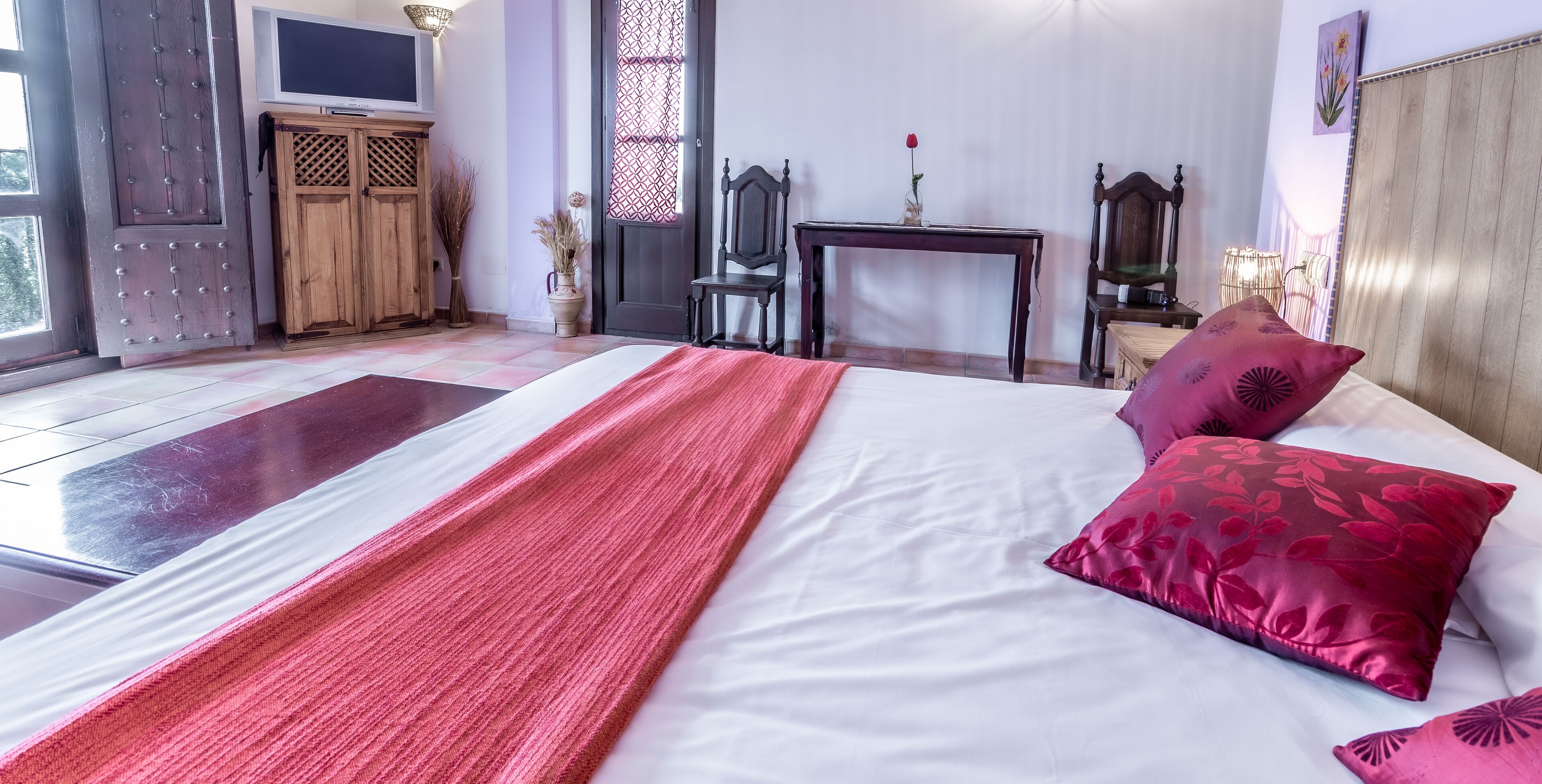 Un dormitorio ordenado con cama, armario de madera, televisor y una mesita con un florero.