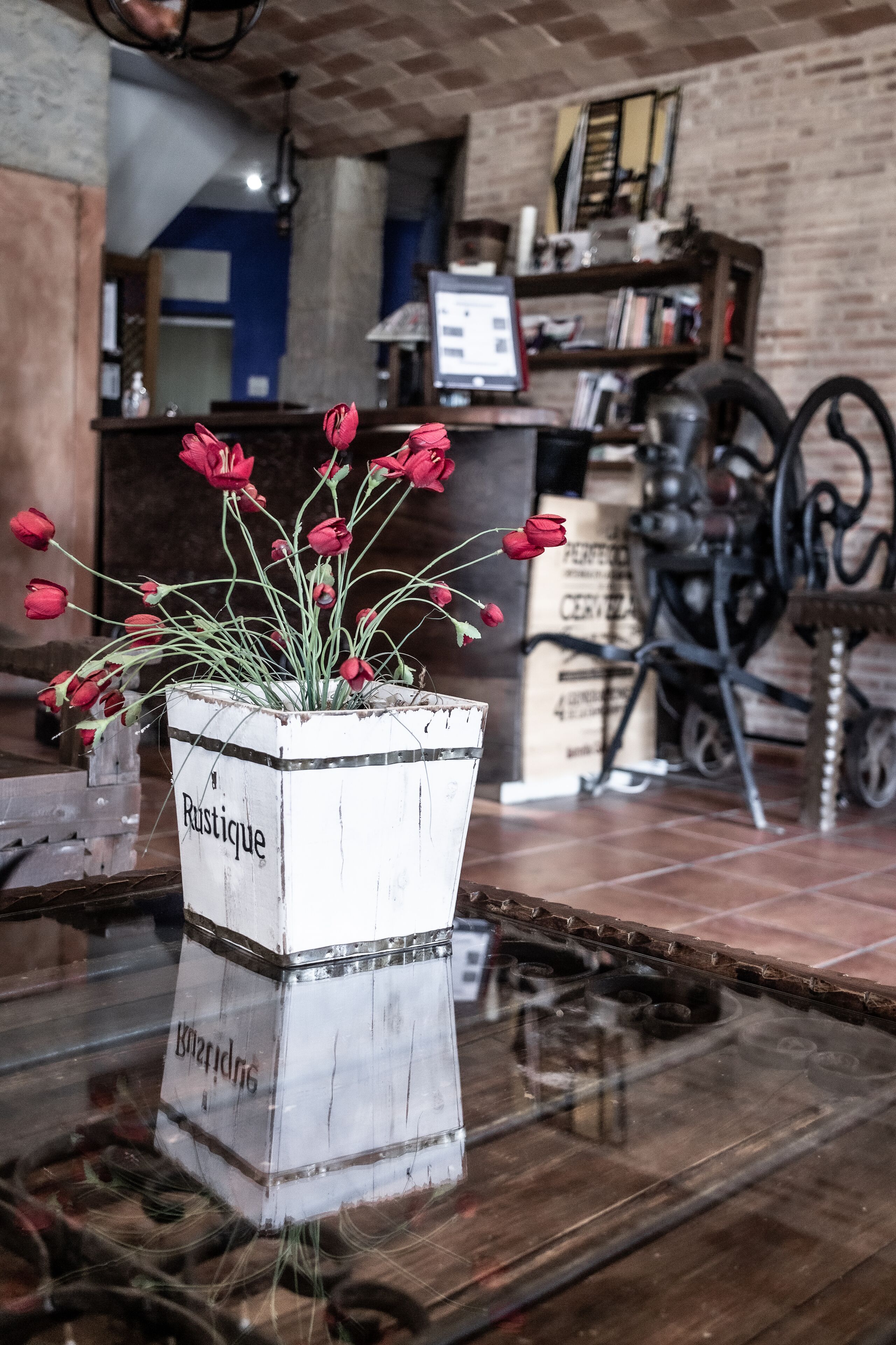 Ein rustikaler Blumentopf mit roten Blüten auf einem Glastisch in einem Raum mit Backsteinwänden und einem Holzregal im Hintergrund.