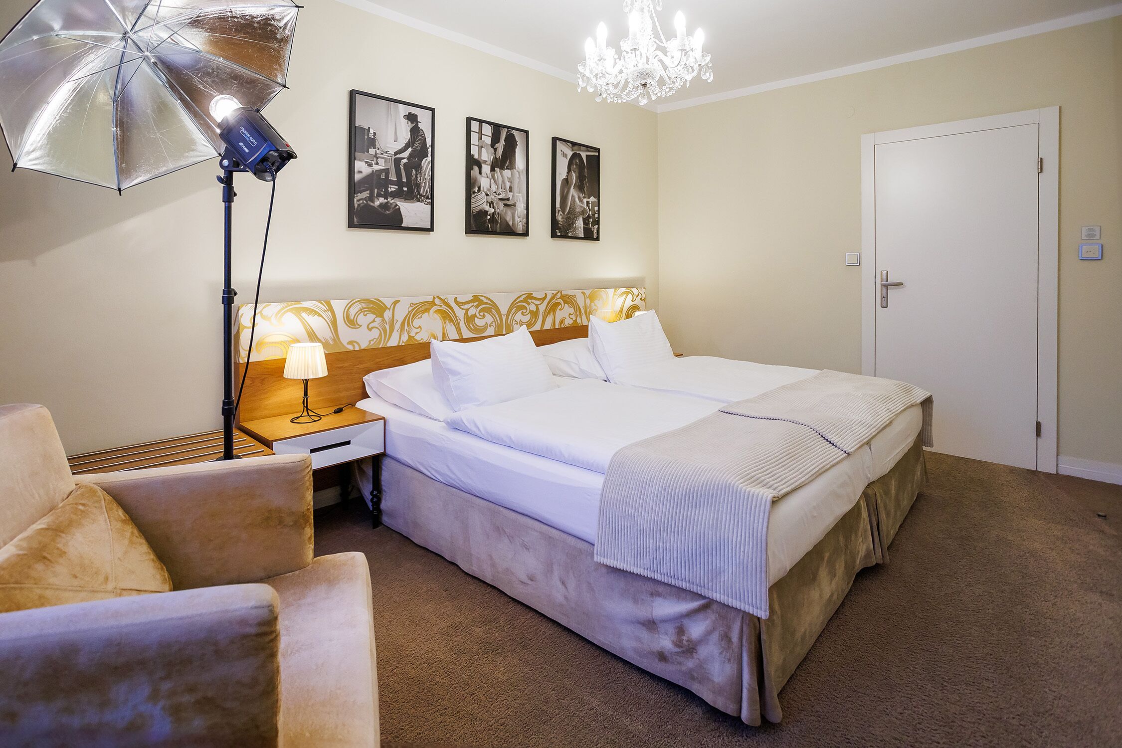Une chambre d’hôtel avec un lit, une table de chevet, une lampe, une chaise, un lustre et trois tableaux encadrés accrochés au mur.
