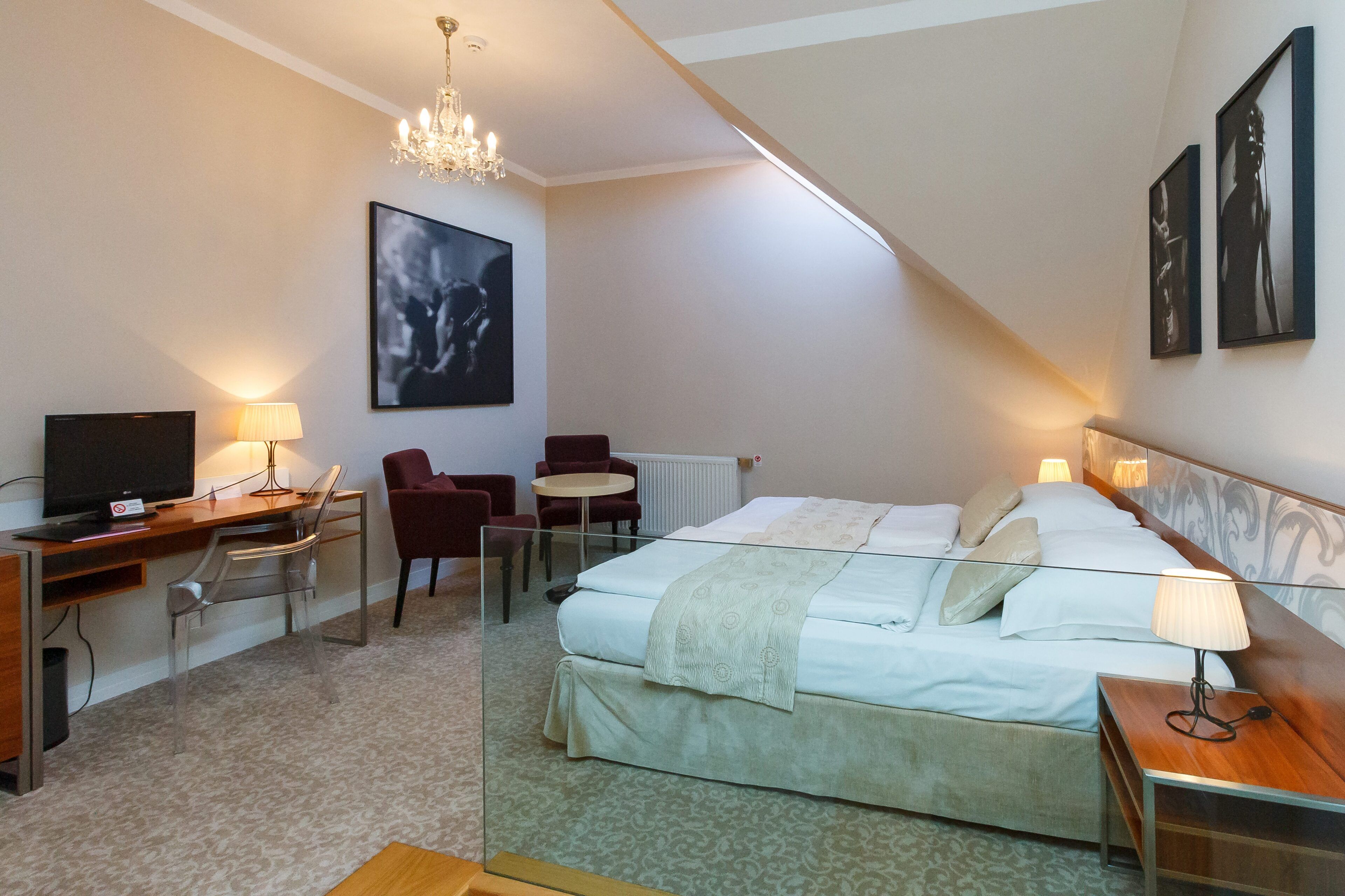 A hotel room with a bed, desk, chair, TV, and artwork on the walls.