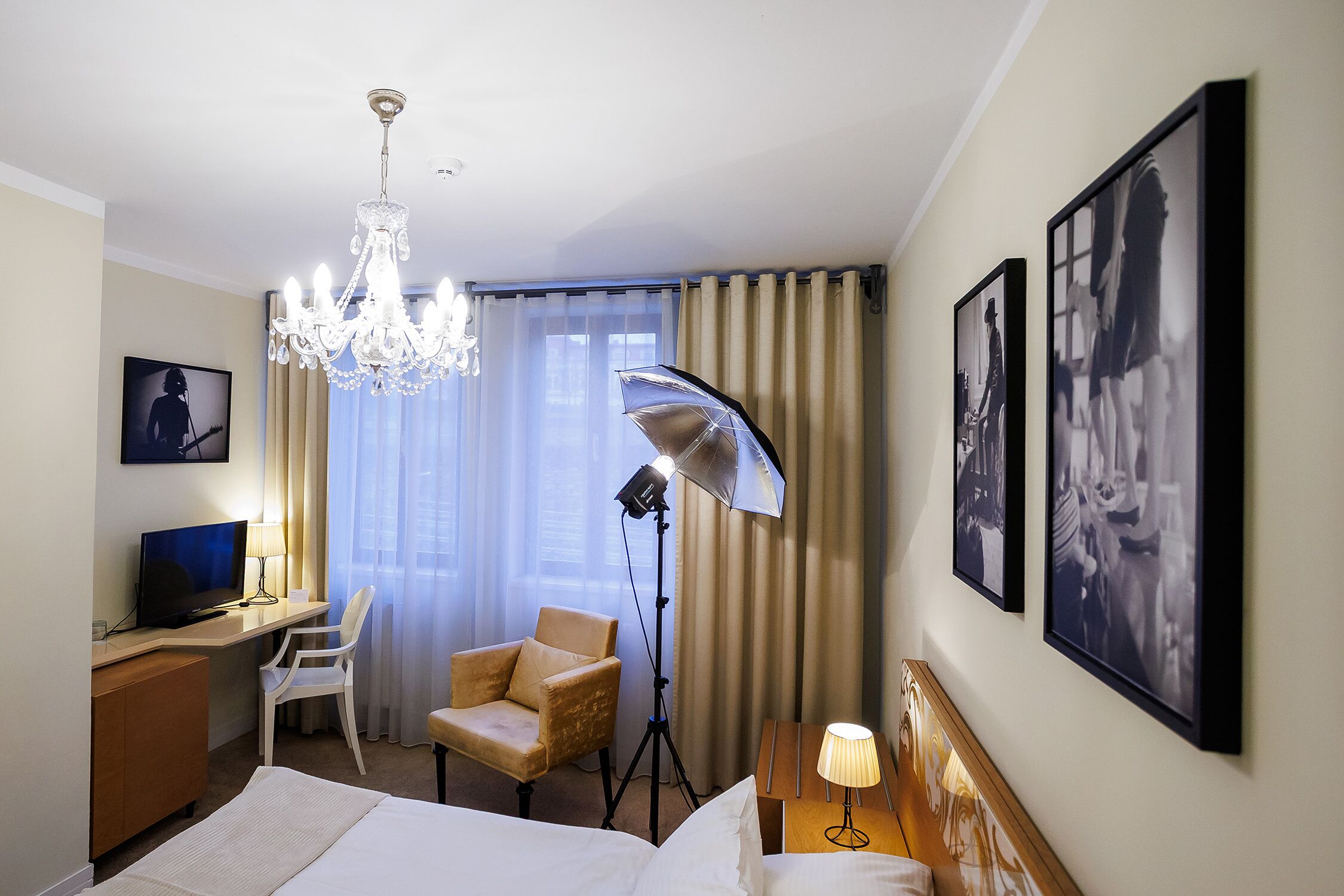 Quarto de hotel com uma cama, uma escrivaninha com computador, uma cadeira, um candelabro e duas quadros na parede.