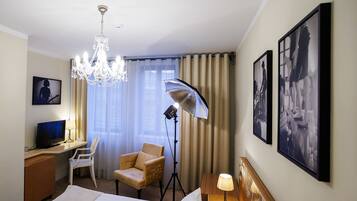 Quarto de hotel com uma cama, uma escrivaninha com computador, uma cadeira, um candelabro e duas quadros na parede.