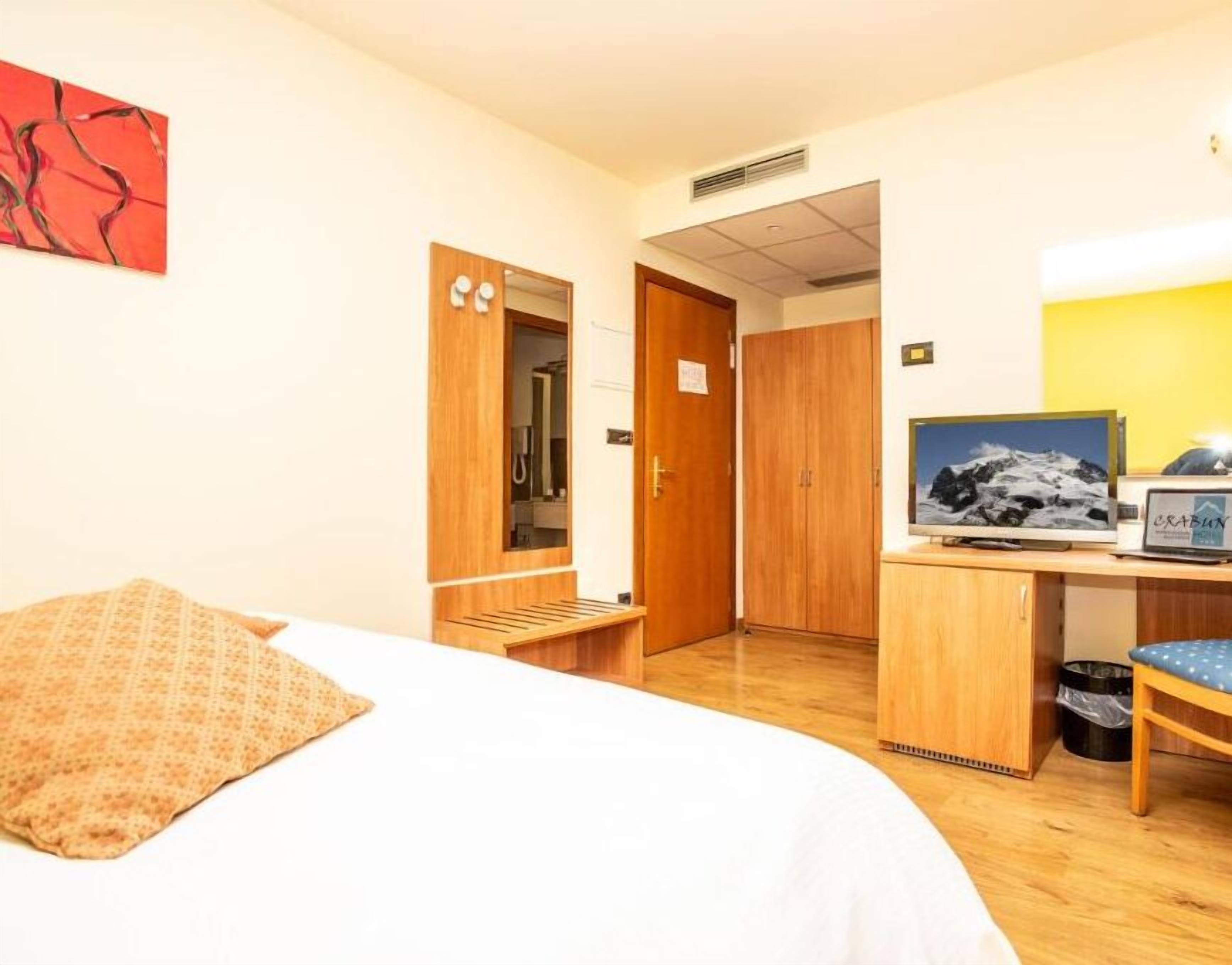 A hotel room with a bed, a desk with a computer and a TV, a wooden wardrobe, and a bathroom visible through an open door.