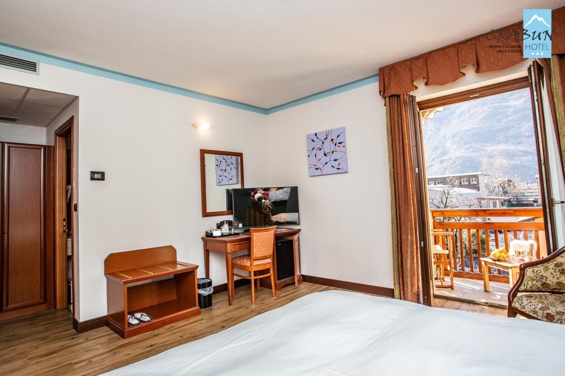 Een hotelkamer met een bed, een bureau met een computer, een stoel en een balkon met uitzicht op de bergen.