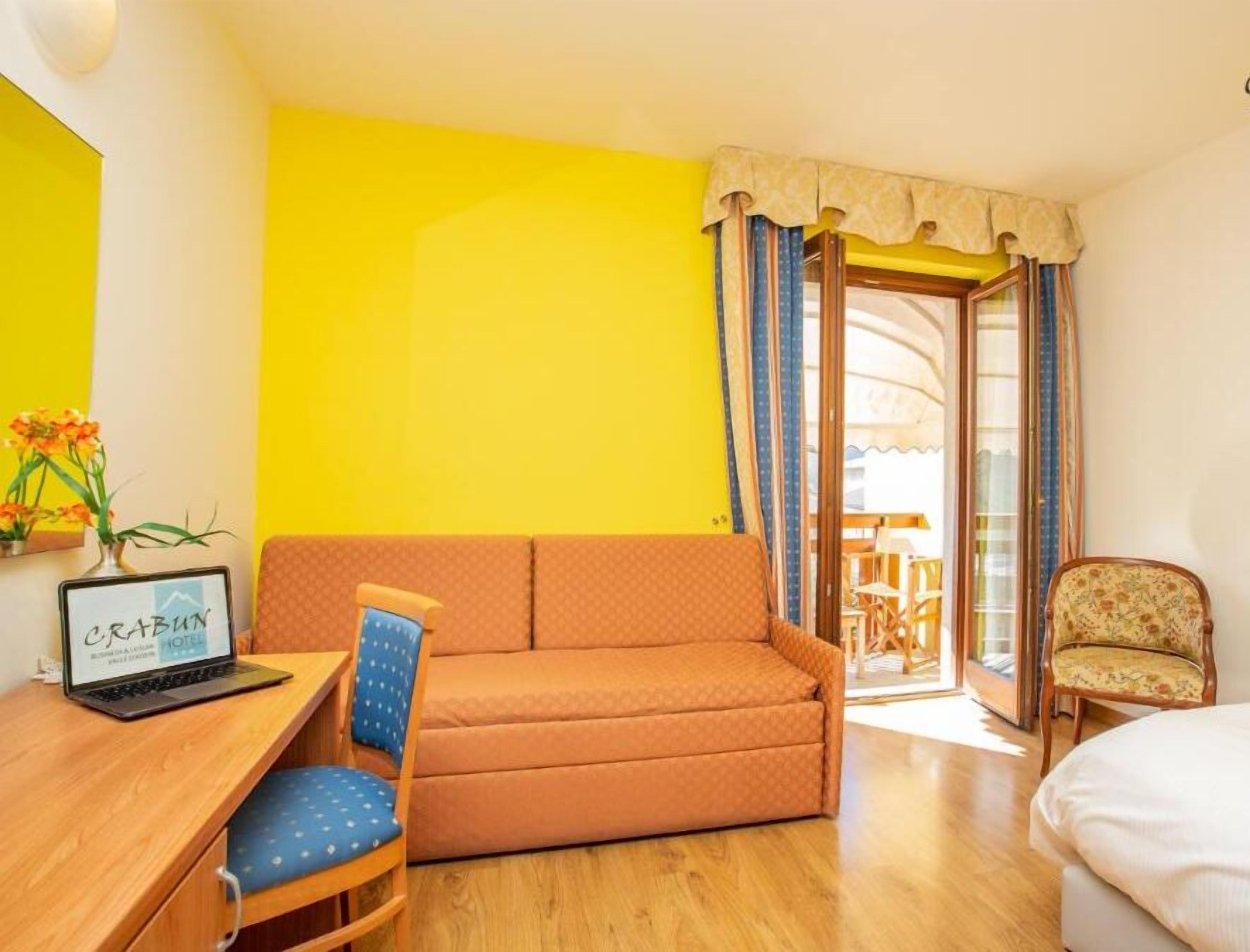 A hotel room with a yellow wall, a wooden desk with a laptop, a blue chair, an orange sofa, and a wooden chair.