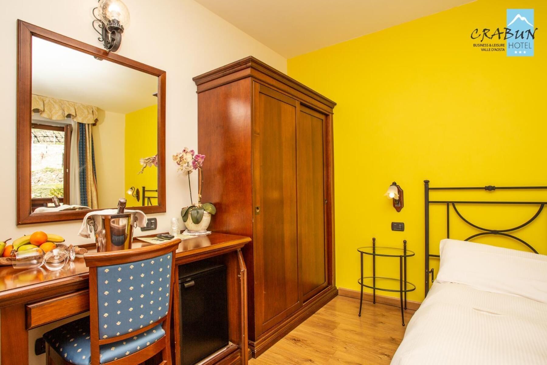 A hotel room with a wooden desk, a chair, a bed, a wardrobe, and a mirror.