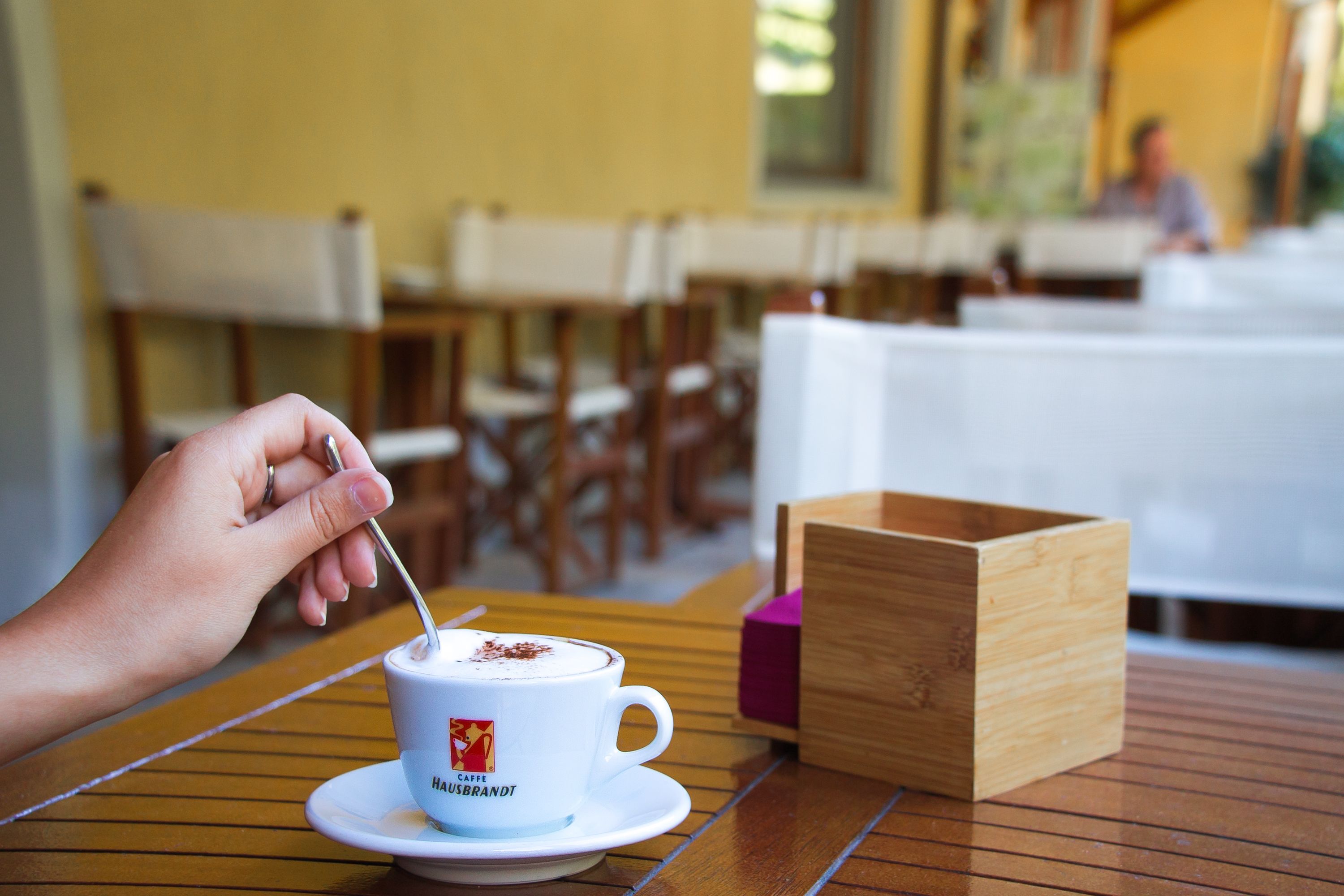 A person stirring a cup of coffee on a wooden table in a cafe setting.