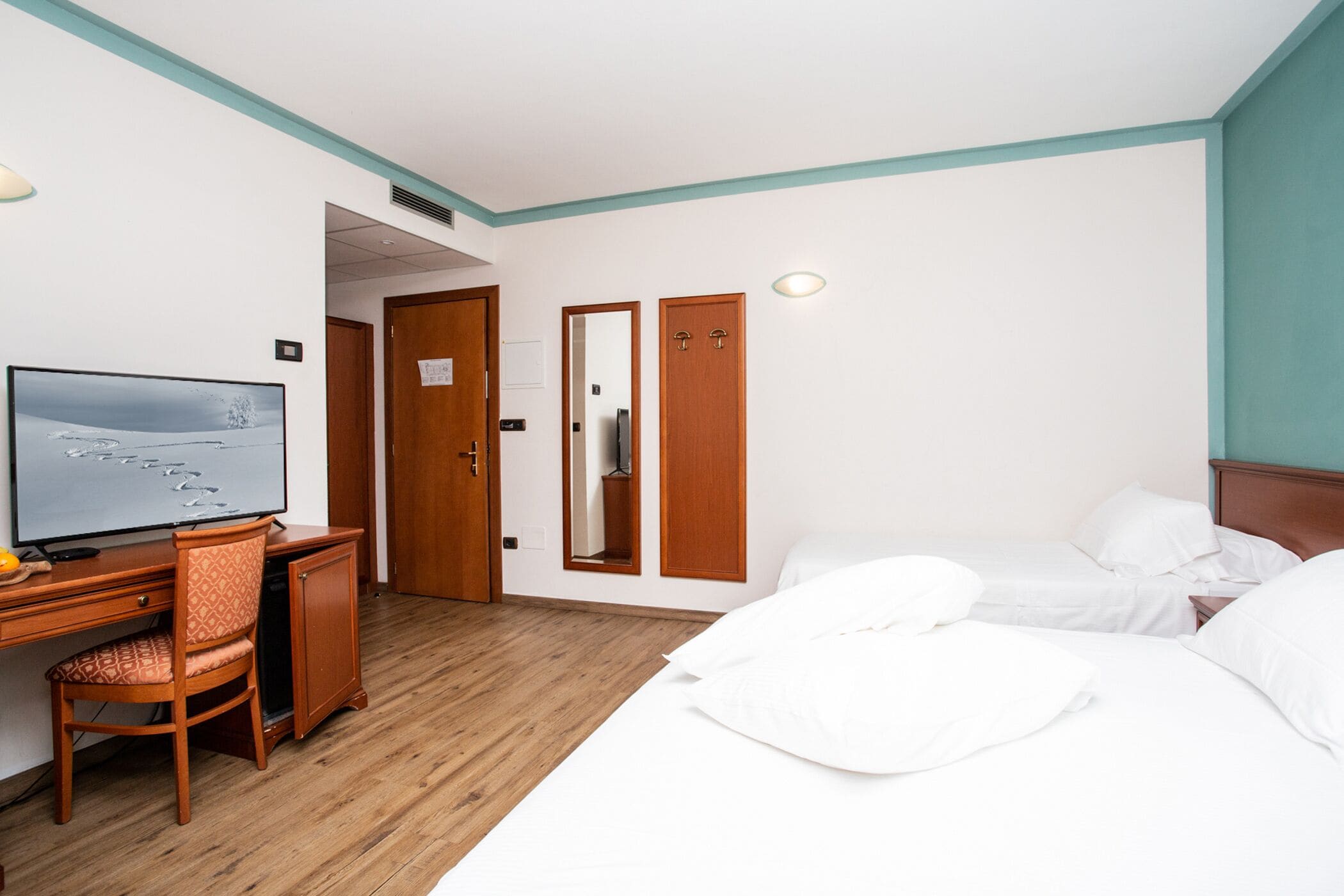 Een hotelkamer met een houten bureau, een televisie, een bed met wit beddengoed en een spiegel.