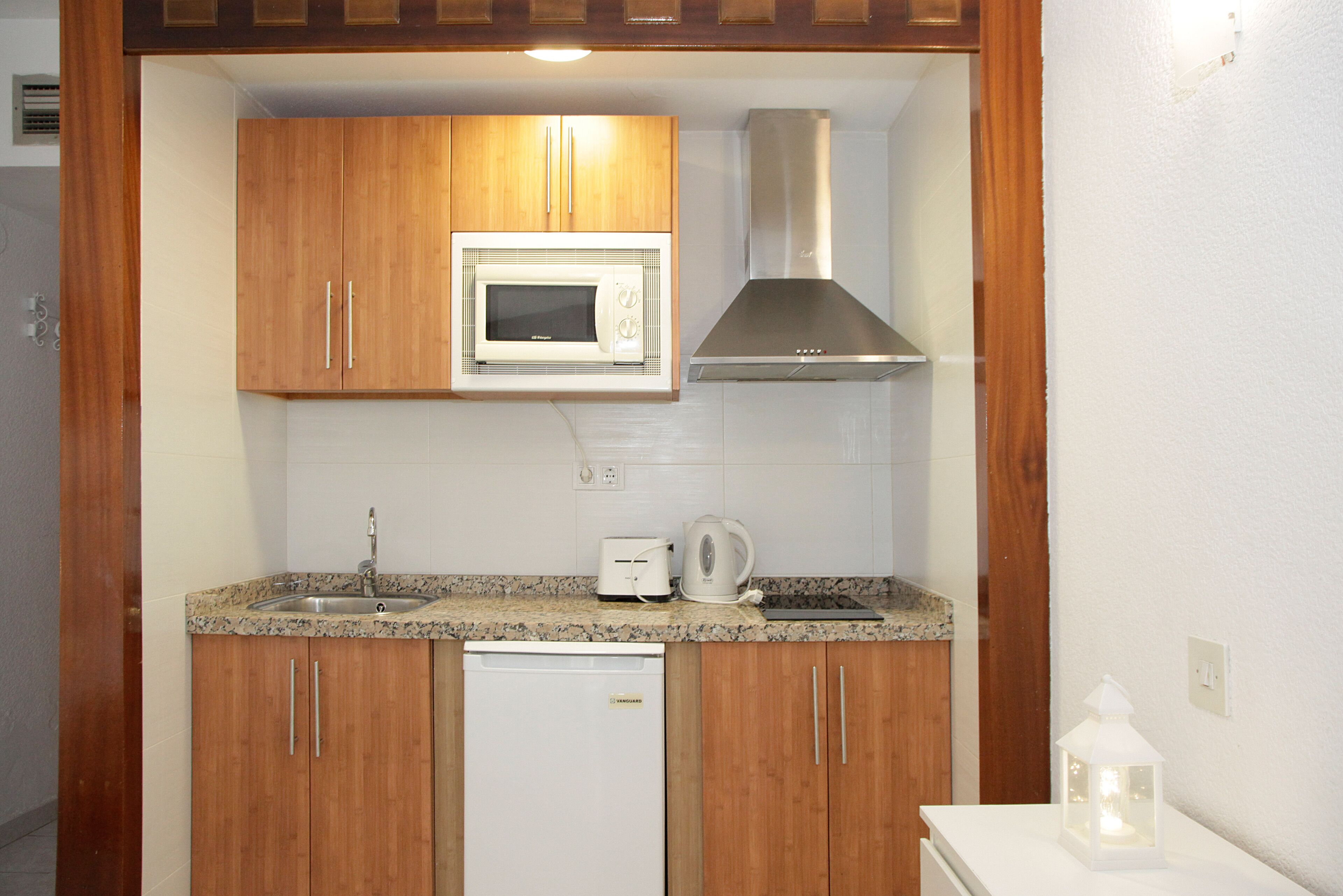 A compact kitchen with wooden cabinets, a built-in microwave, a stainless steel range hood, a dishwasher, and a countertop with a kettle and toaster.