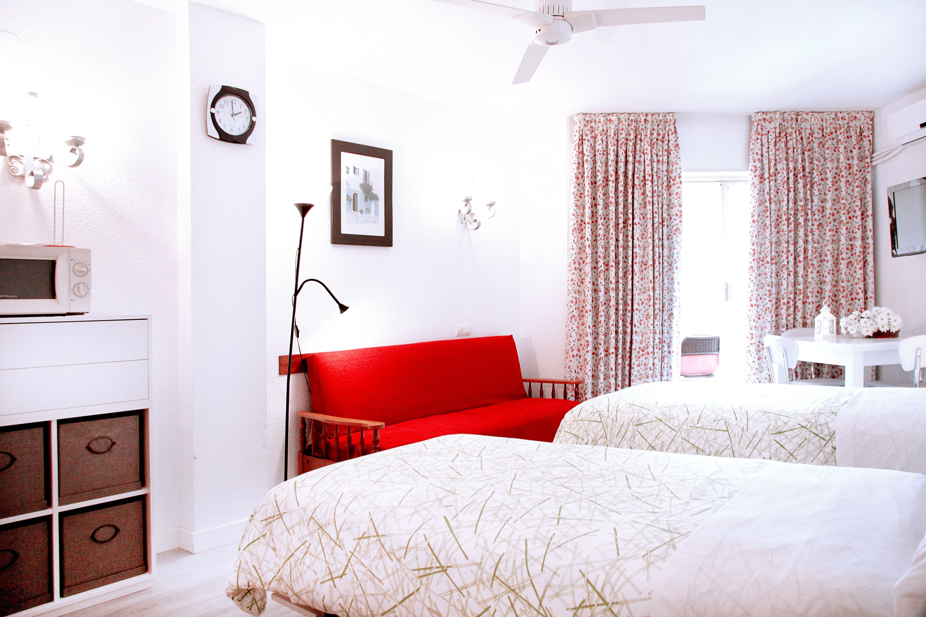 A hotel room with a red sofa, two beds with patterned bedding, a white desk with a chair, a clock on the wall, and a ceiling fan.