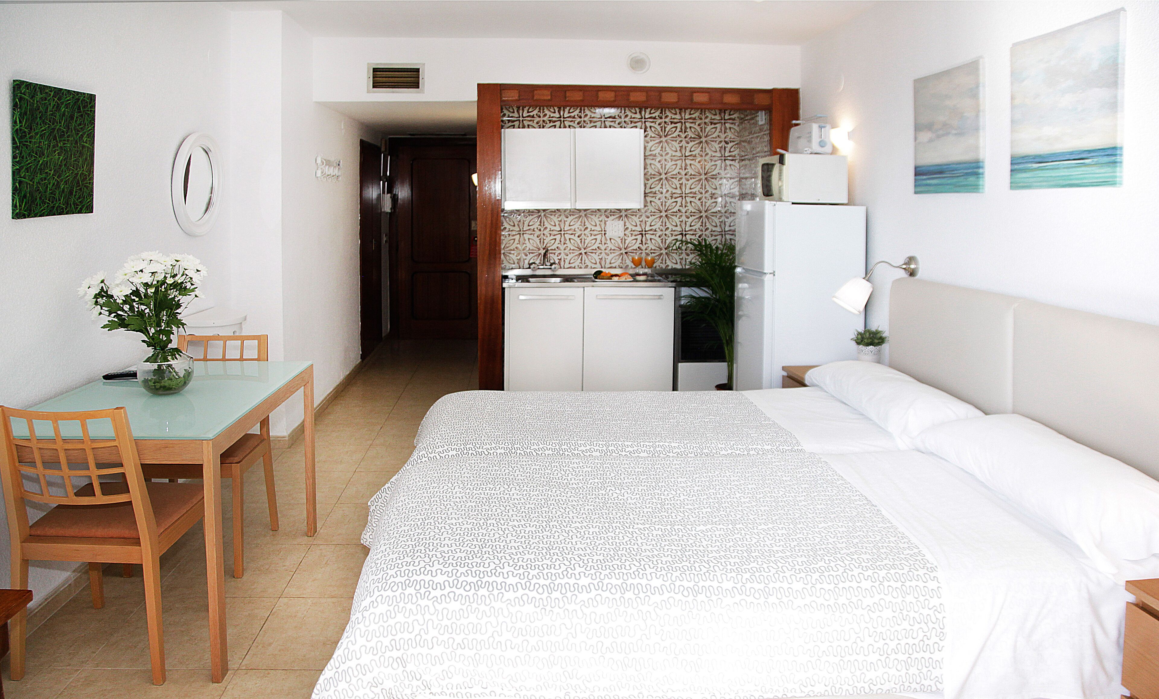 Quarto de hotel com cama, mesa de jantar com cadeiras, cozinha compacta e banheiro visível por uma porta aberta.