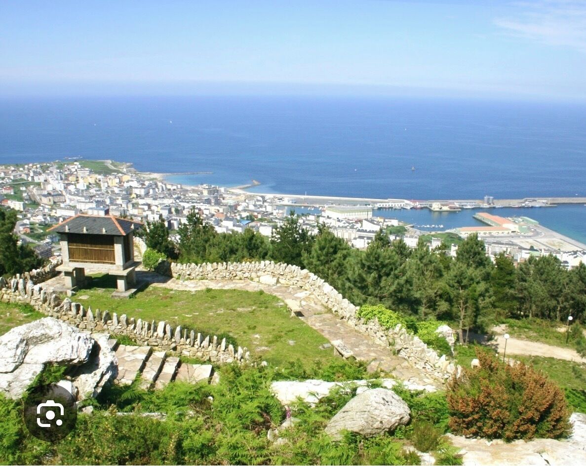 A coastal town with a historic stone wall, a small traditional building, and a view of the sea and a harbor.