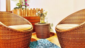 A wicker chair with a cushion, a small round table with a vase of flowers, and a wooden plant stand with a potted plant. - Hotel Galatea (Burela)