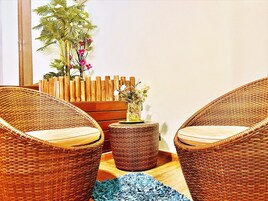 Una silla de mimbre con un cojín, una mesita redonda con un florero y un atril de madera con una planta en maceta.