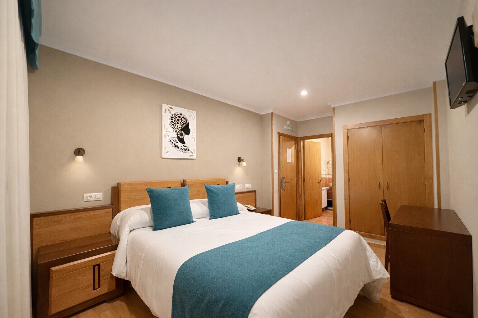 Un dormitorio con cama, mesitas de noche, armario, televisor de pared y puerta que da a otra habitación.