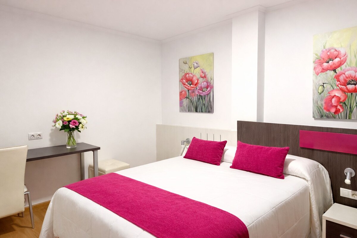 A hotel room with a bed, a desk, a chair, and two paintings on the wall.