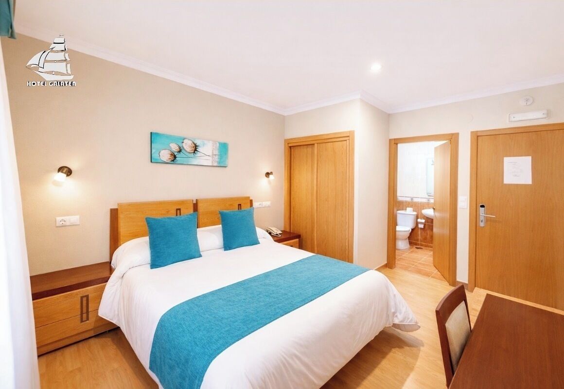 A hotel room with a bed, bedside tables, a nightstand, a painting, a wooden door, and a bathroom visible through an open door.