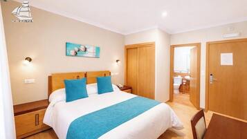 Habitación de hotel con cama, mesitas de noche, un cuadro, una puerta de madera y un baño visible a través de una puerta abierta.
