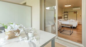 A modern hotel room with a marble table, a small white vase with flowers, a bed with white linens, a wooden floor, and a glass partition.
