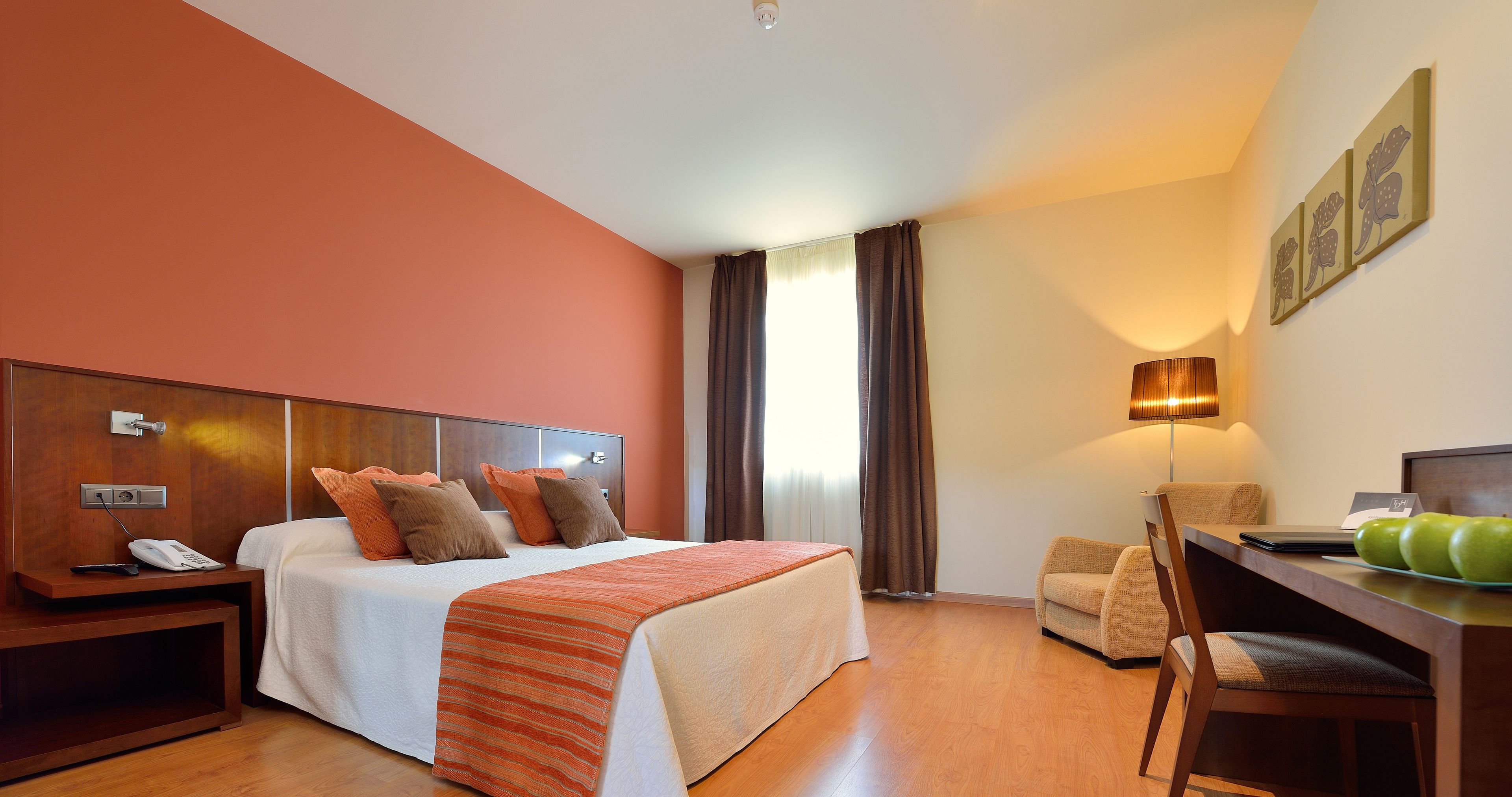 A hotel room with a bed, bedside table, lamp, chair, and a desk with apples.