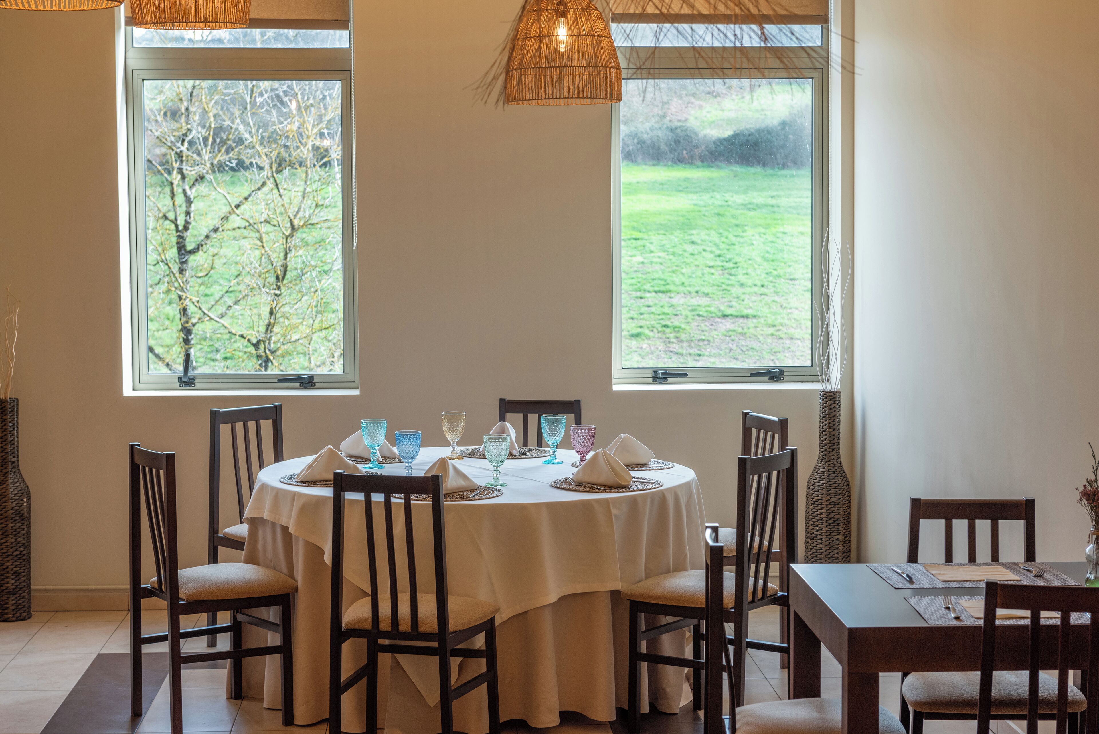 Un coin repas avec une table ronde dressée pour quatre personnes, entourée de chaises, et deux fenêtres donnant sur un champ verdoyant.