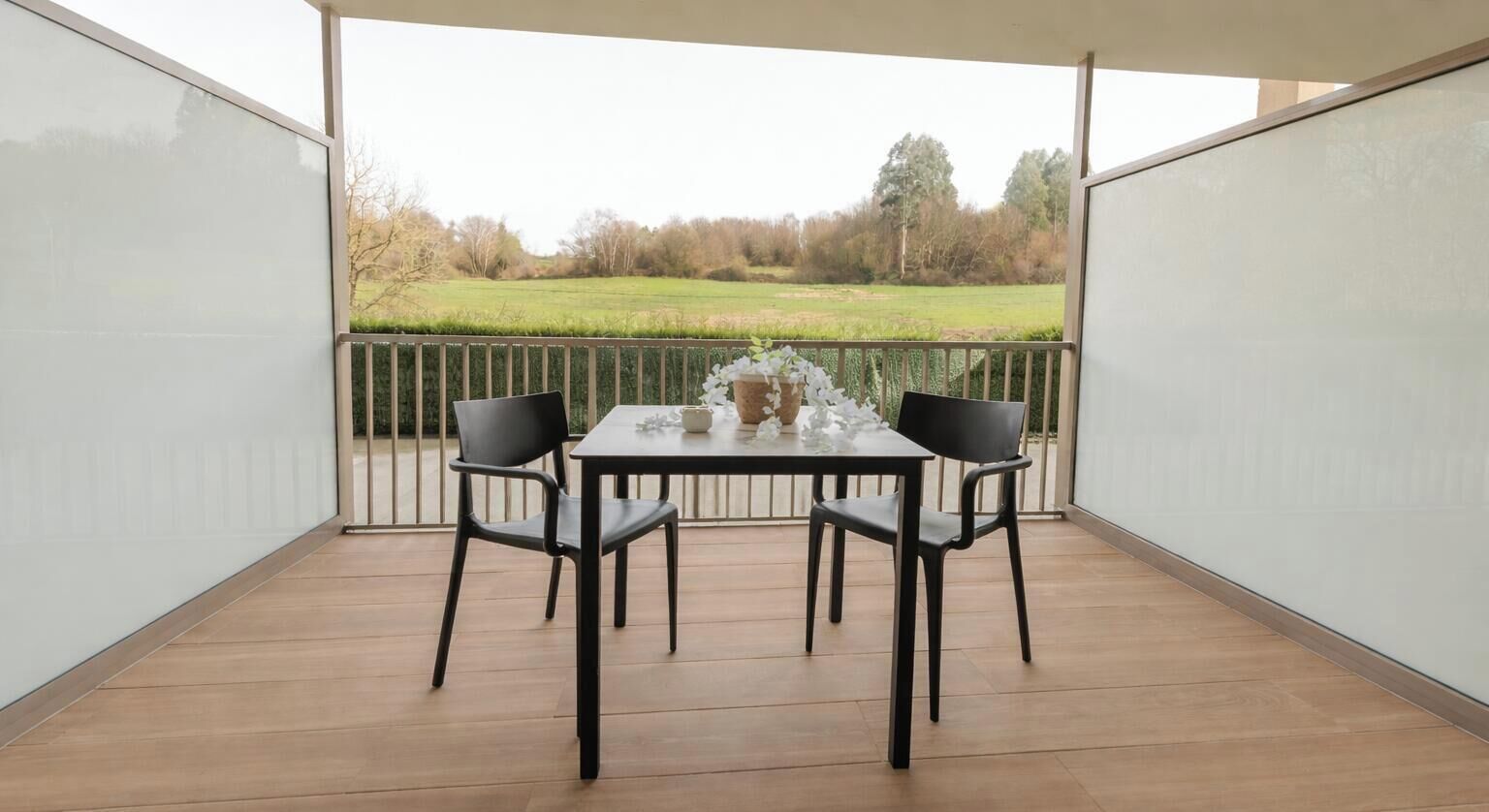 A balcony with a table and chairs, a vase with flowers, and a view of a green field.