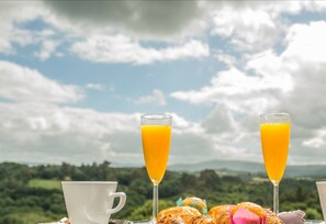 A table with two glasses of orange juice, a white mug, and pastries against a scenic outdoor backdrop.