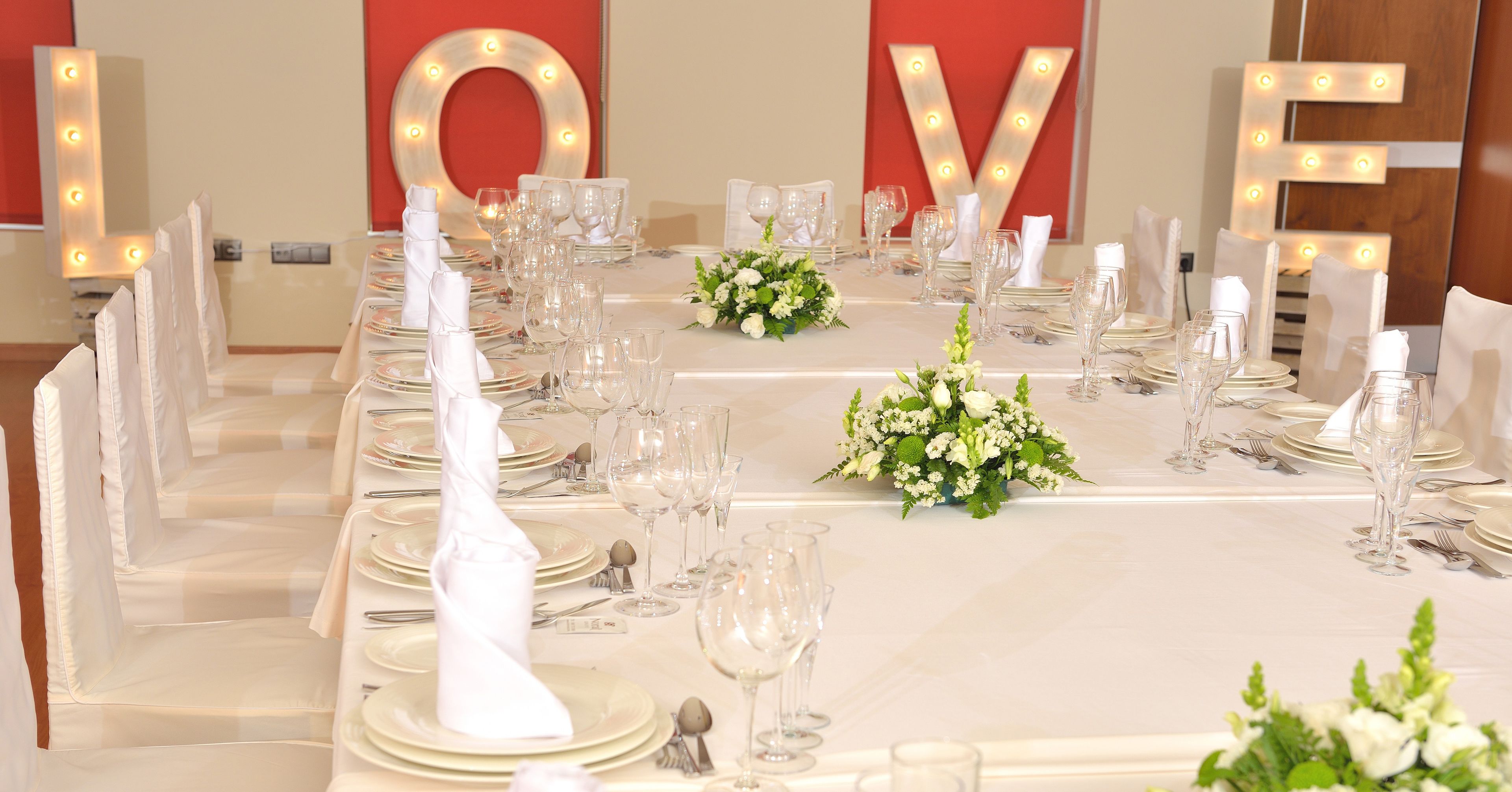 A banquet table set with white tablecloths, plates, glasses, and cutlery, with illuminated 'LOVE' letters in the background.