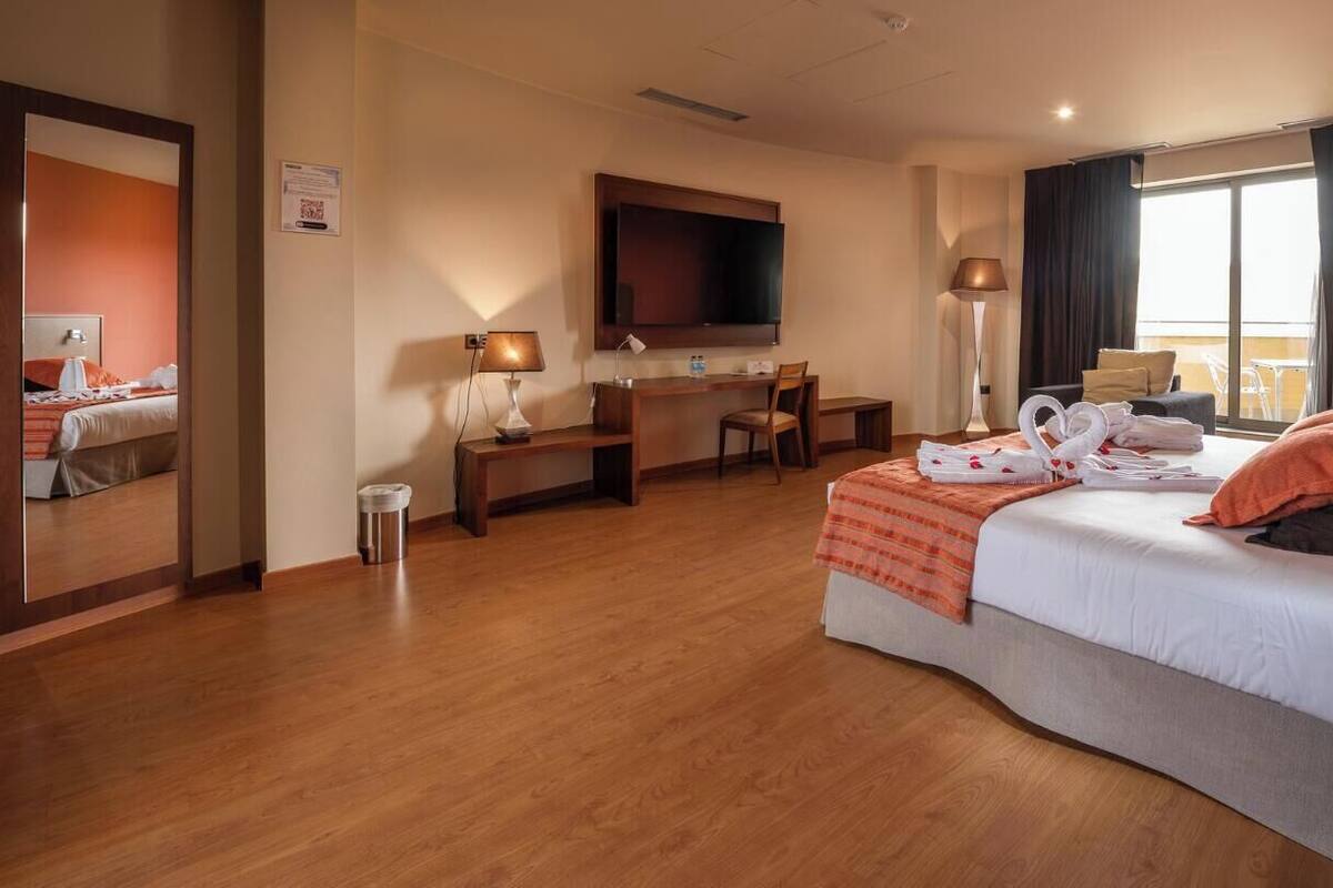 Habitación de hotel con cama, escritorio, silla, televisor y balcón.
