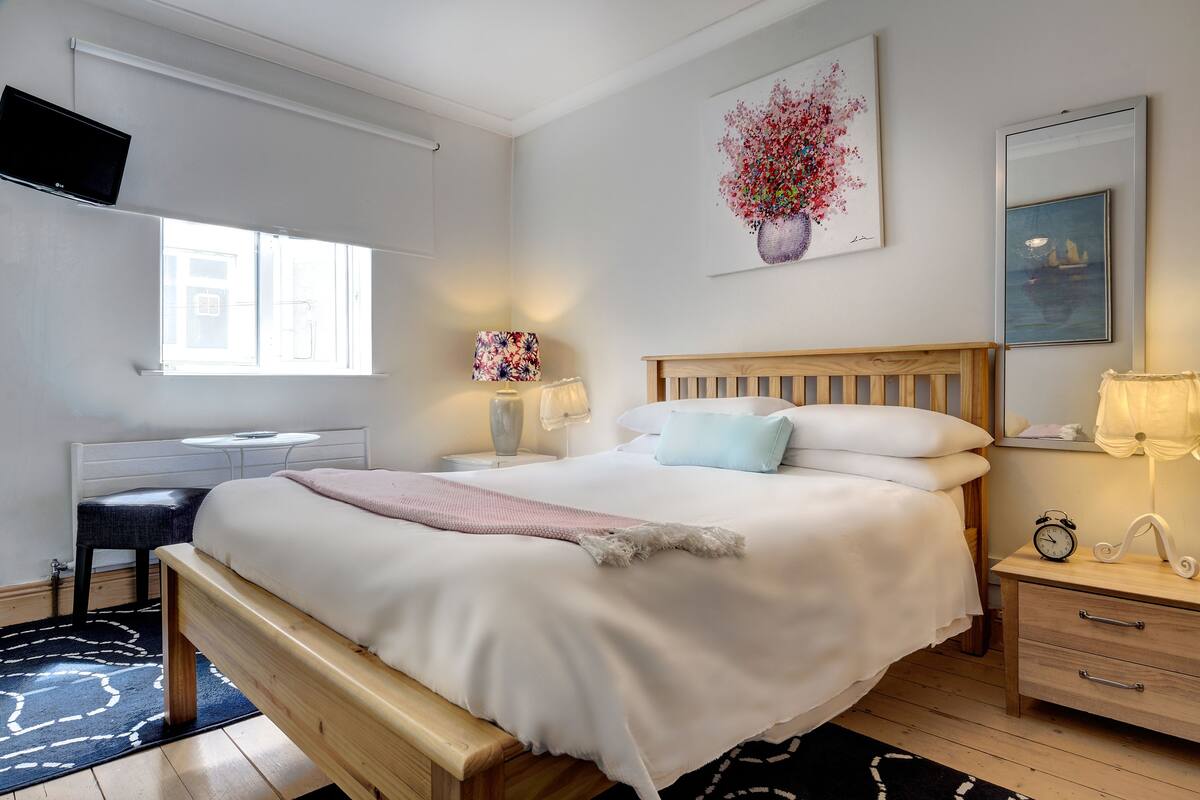 A bedroom with a wooden bed, white bedding, a desk with a chair, a TV, and artwork on the walls.