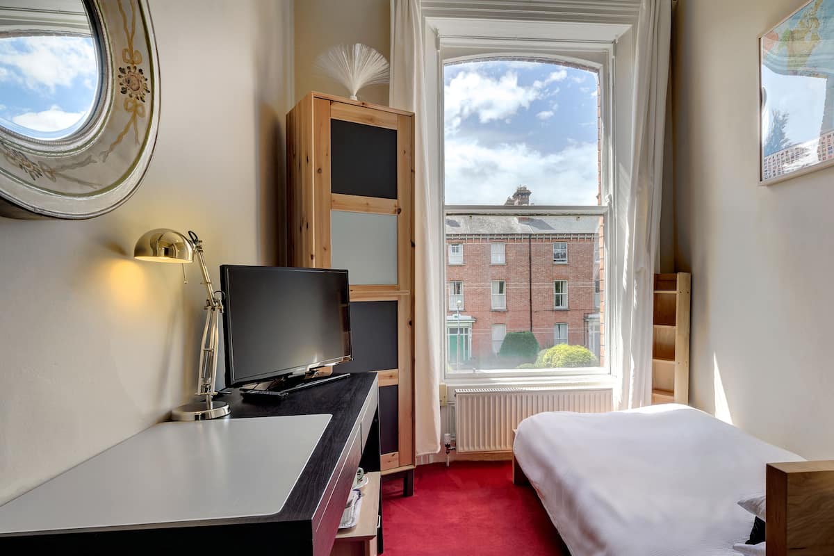 A hotel room with a bed, a desk with a computer, a television, a window with a view of buildings, and a radiator.