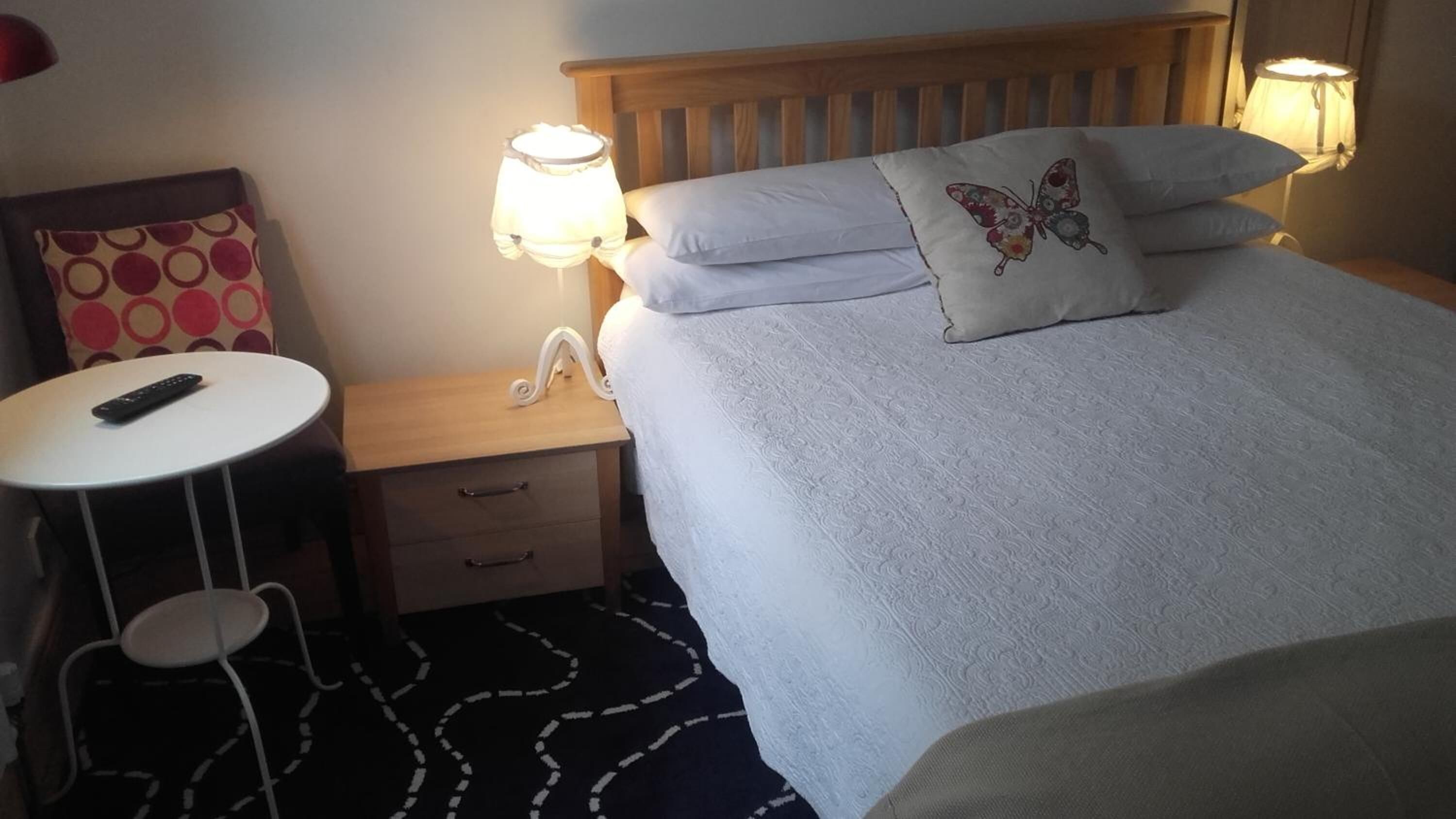Habitación de hotel con una cama, lámparas de noche, una mesita con un control remoto, una silla y una alfombra estampada.
