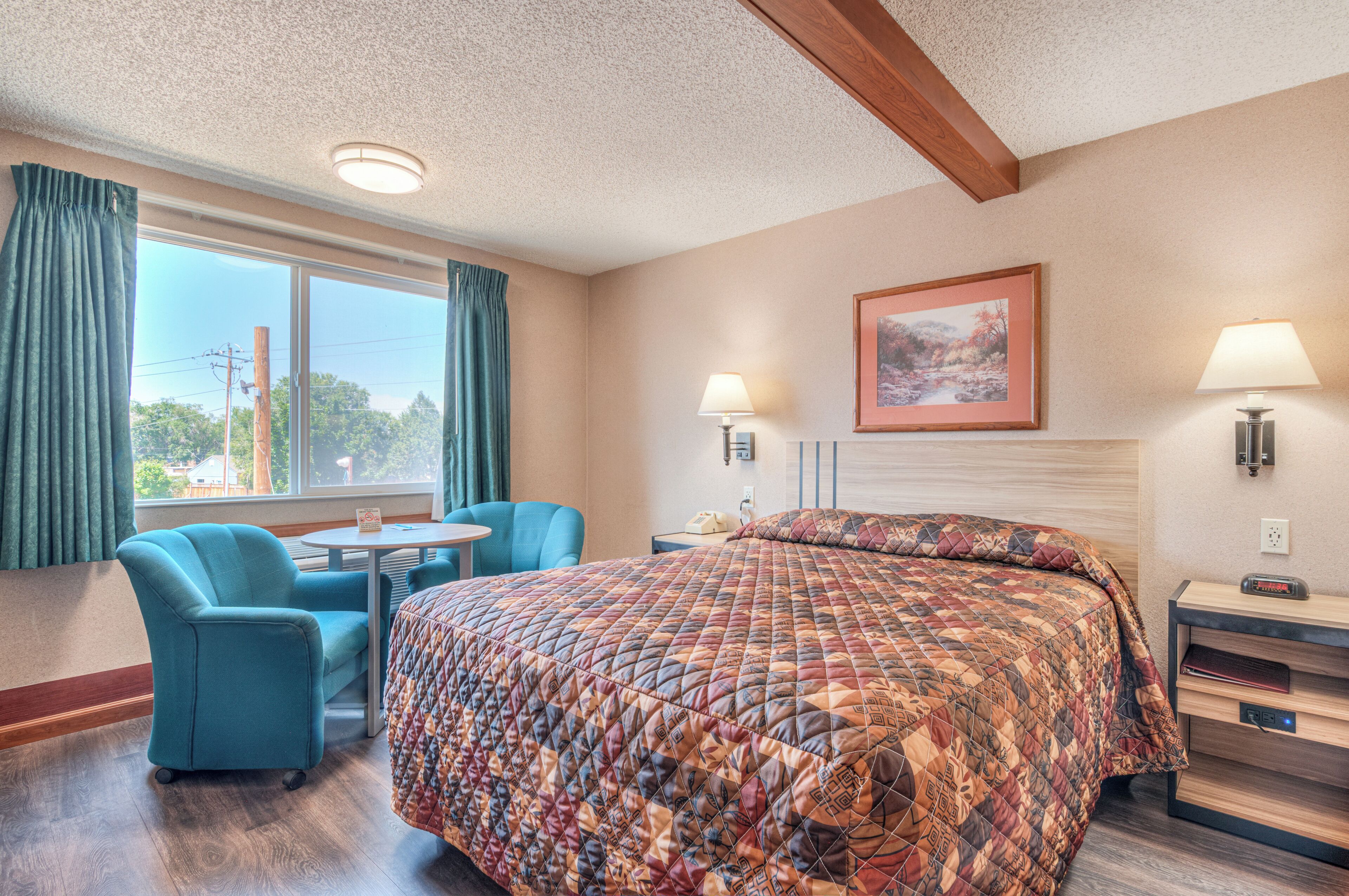 A hotel room with a bed, a desk, a chair, a window with curtains, and a painting on the wall.