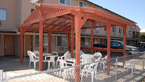 A covered outdoor seating area with white plastic chairs and tables, a red wooden pergola, and a chain-link fence.