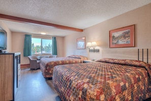 A hotel room with two beds, a desk, a chair, a window with curtains, a painting on the wall, and a lamp.