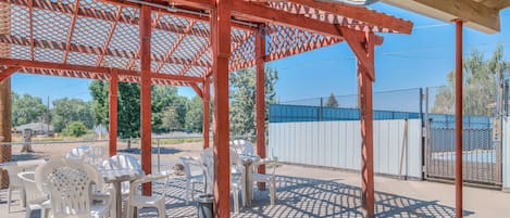 A covered outdoor seating area with tables and chairs, a pergola structure, and a fenced area in the background.