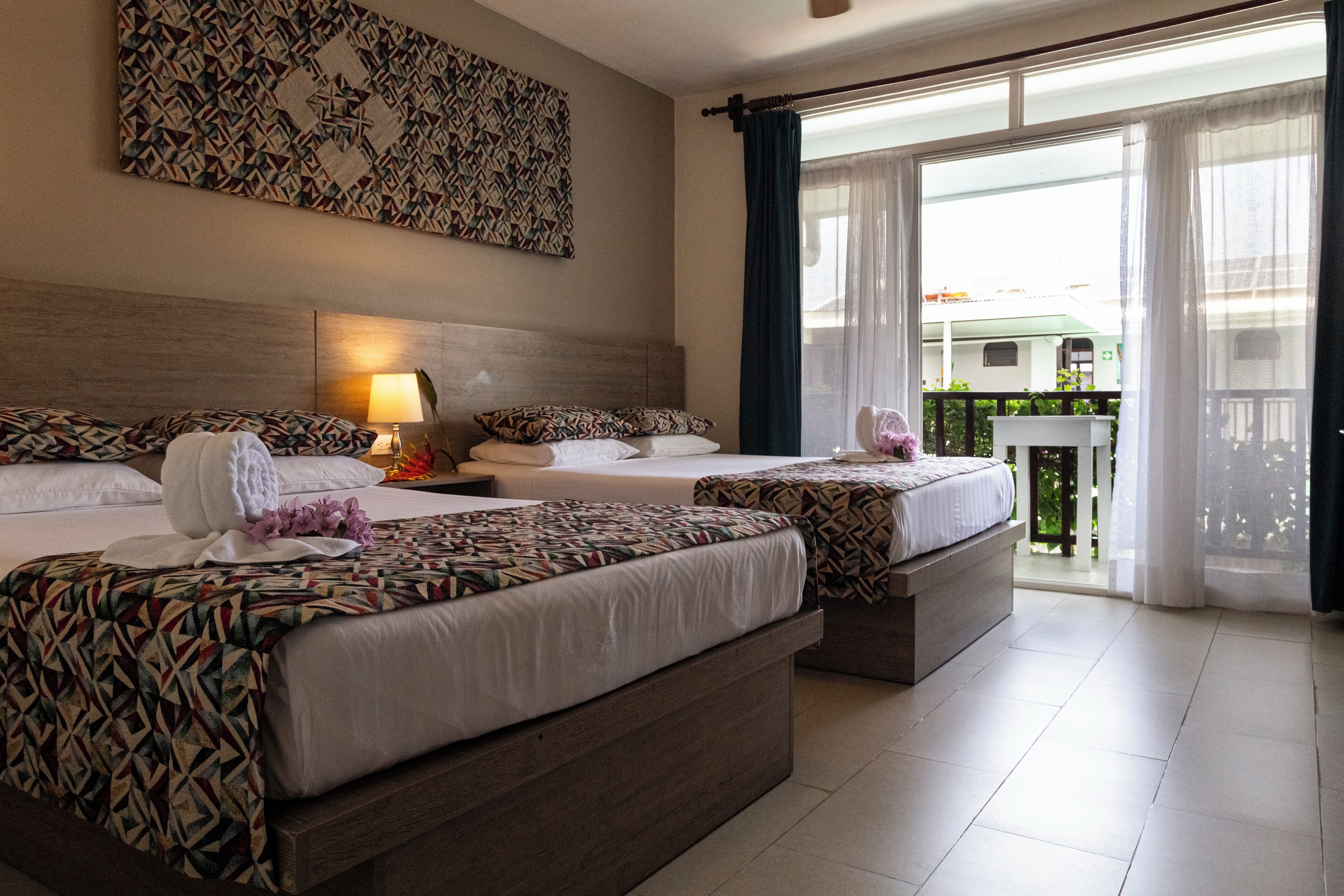 A hotel room with two beds, a large window with curtains, a wall-mounted artwork, and a view of the outside balcony.