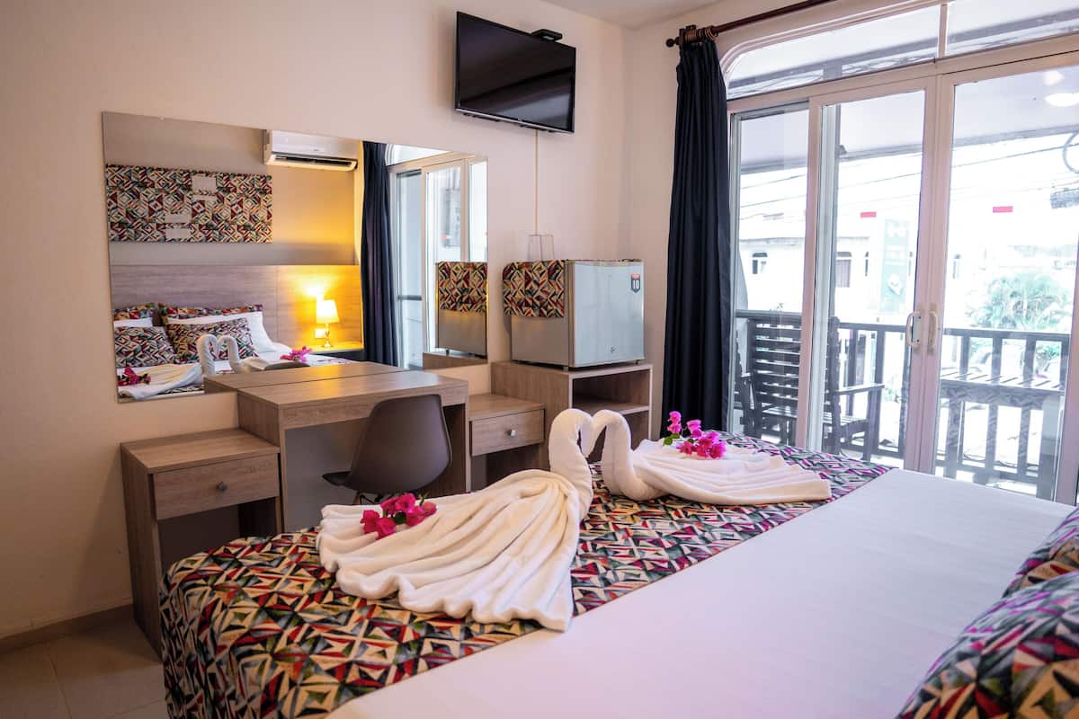 Una habitación de hotel con cama, escritorio, silla, televisor y balcón con barandilla.