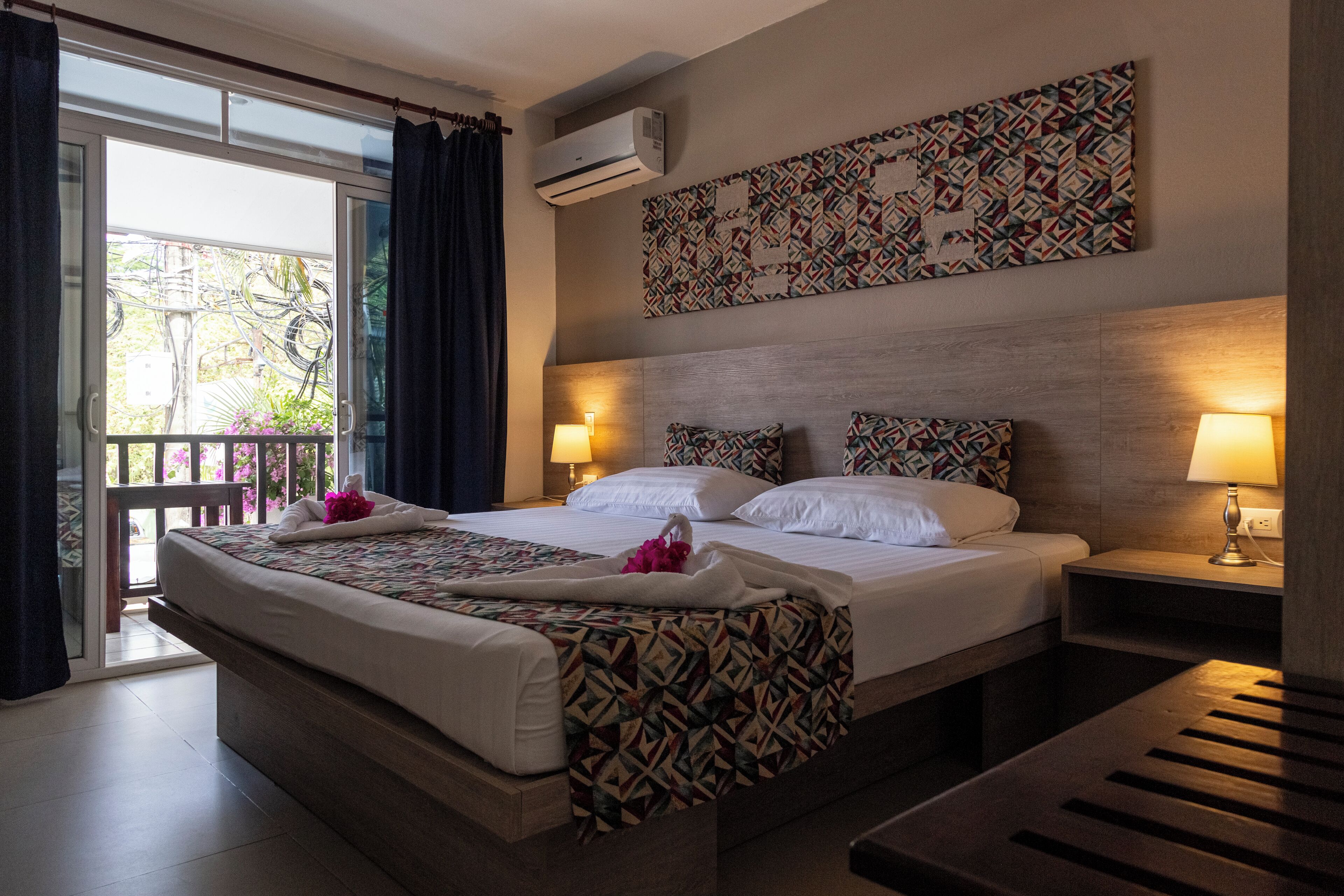 A double bed with patterned bedding, two bedside lamps, a wall-mounted air conditioner, and a balcony with curtains.