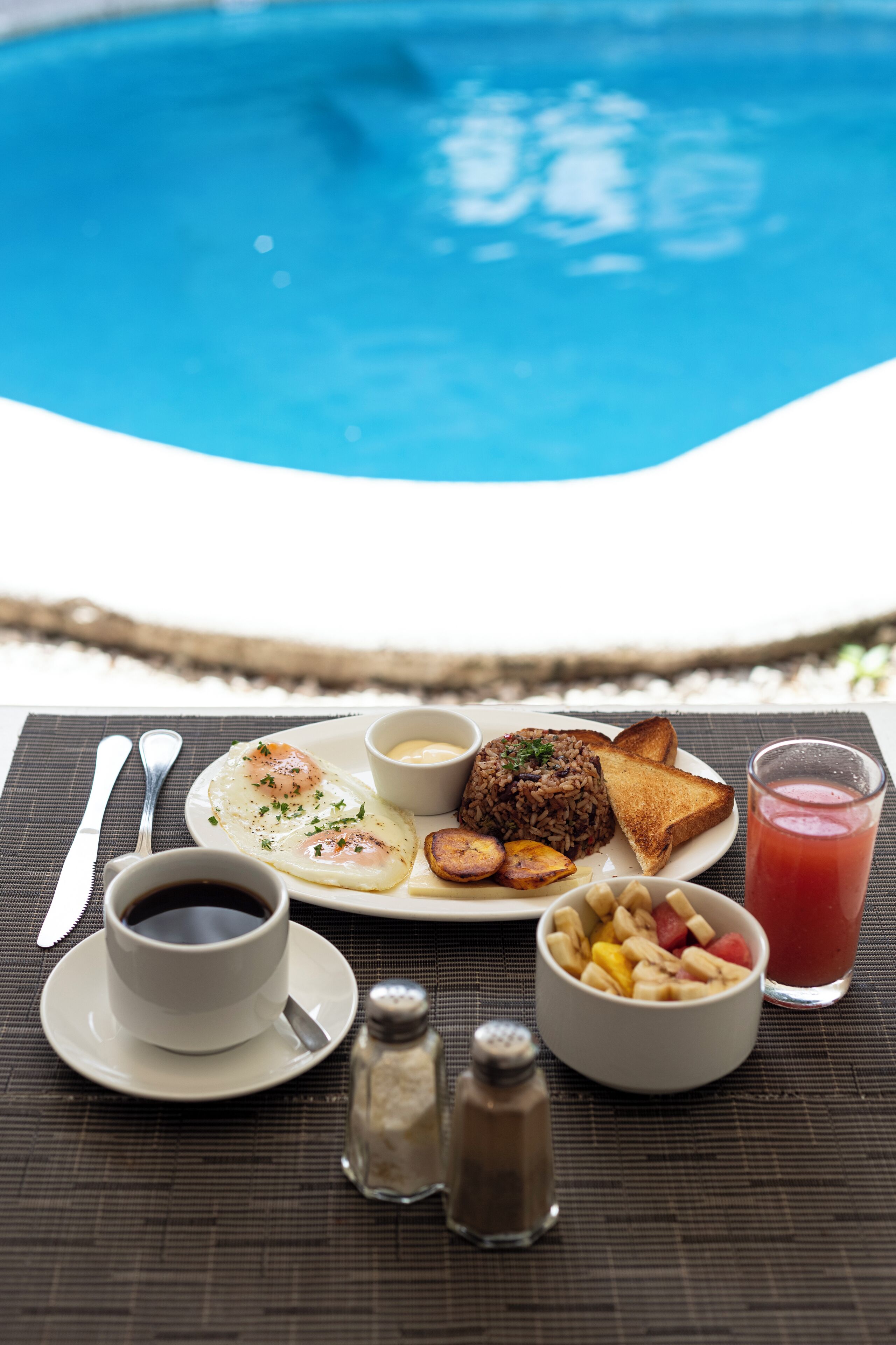 A breakfast setting by a pool with toast, eggs, fruit, coffee, and a drink.