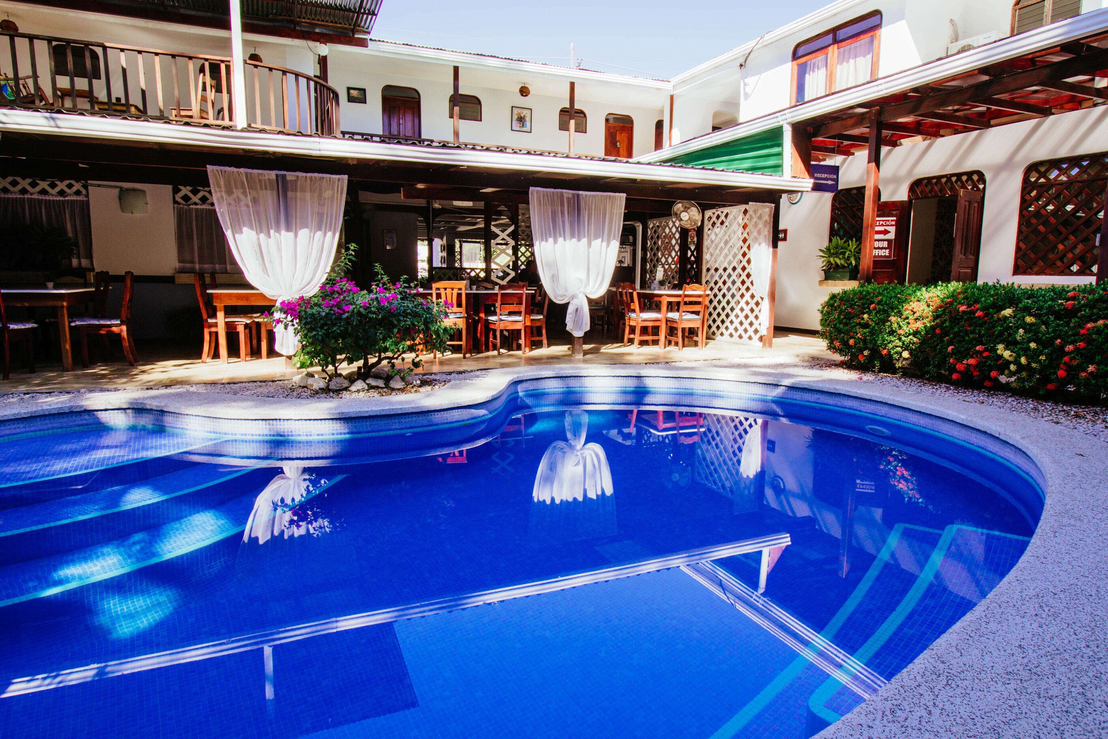 A swimming pool with a clear blue water, surrounded by wooden chairs and tables, and a covered seating area with white curtains.