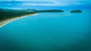 A coastal landscape with a beach, clear blue water, and lush green hills in the background.