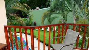 A balcony with a wooden railing, a metal chair, and a view of a swimming pool surrounded by greenery.
