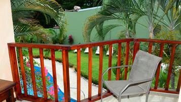 A balcony with a wooden railing, a metal chair, and a view of a swimming pool surrounded by greenery.