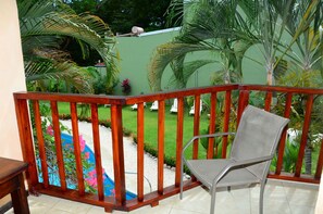 A balcony with a wooden railing, a metal chair, and a view of a swimming pool surrounded by greenery.