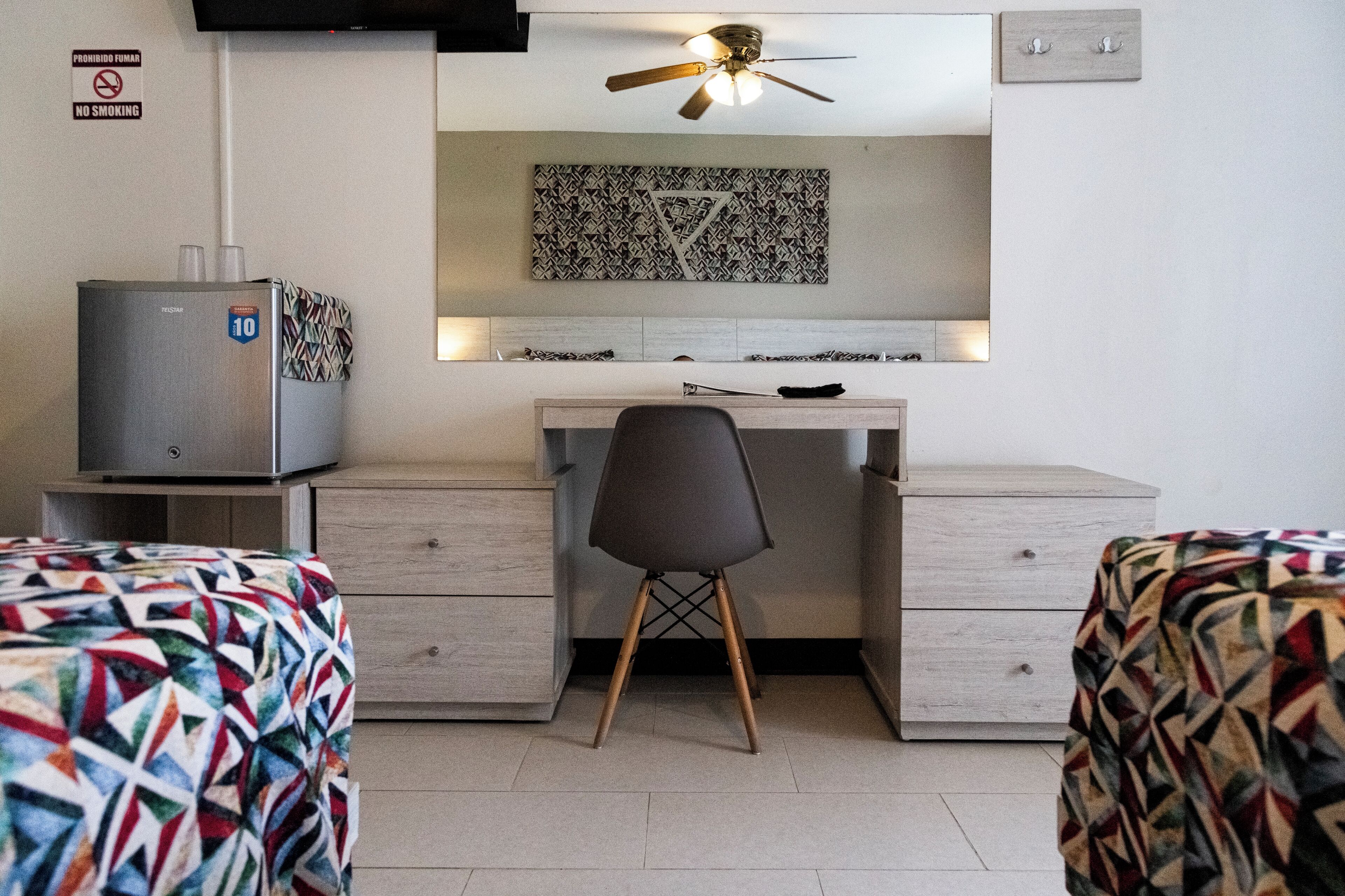 A hotel room with a bed, a desk, a chair, a ceiling fan, a refrigerator, and a wall-mounted artwork.