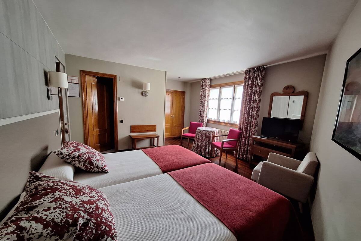 Una habitación de hotel con cama, escritorio, silla, televisor y ventana con cortinas.