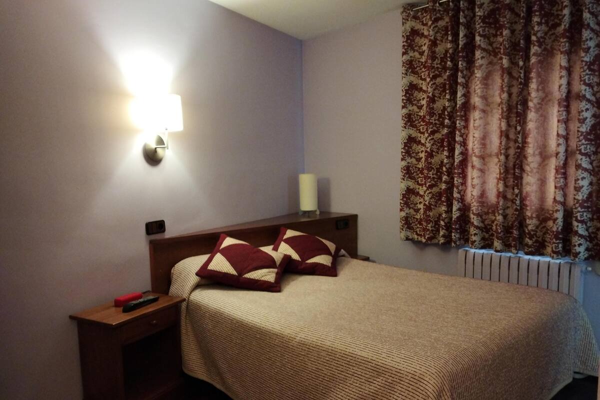 Una cama bien hecha con cabecera, una mesita de noche con lámpara y una ventana con cortinas estampadas.