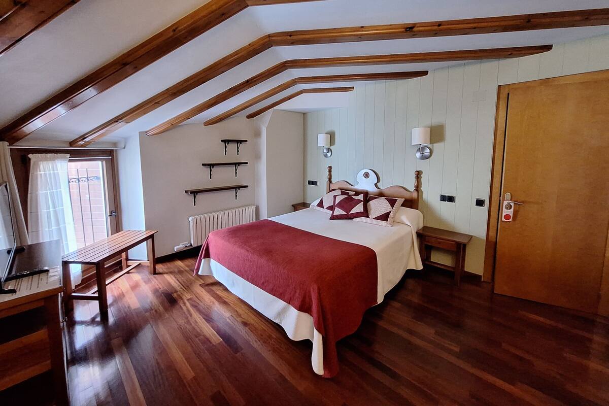 Un dormitorio con techo de madera, una cama con colcha roja, mesitas de noche, un radiador y una puerta con cerradura.