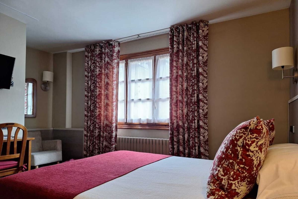 Habitación de hotel con cama, una silla, una mesa y un ventanal con cortinas.
